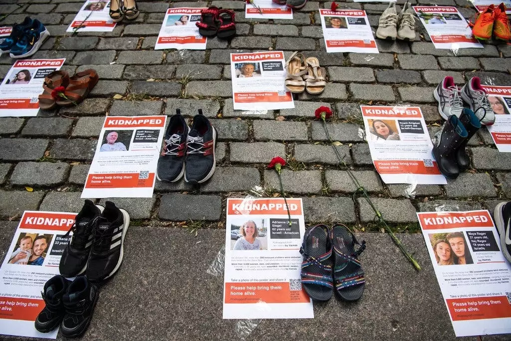 Листовки с фотографиями людей, похищенных 7 октября, разложены на земле перед зданием Организации Объединенных Наций.  Рядом с листовками лежат пары обуви и цветы.